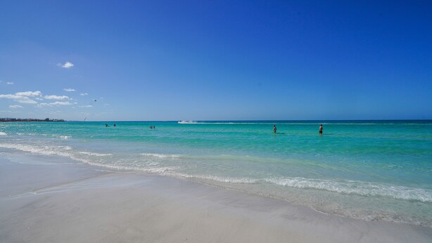 A beach with clear turquoise water, a sandy shore, and a few people in the distance.