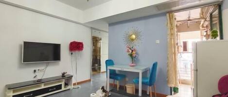 A modern living room with a flat-screen TV, a coffee table, a sofa, a dining table with blue chairs, and a decorative mirror.