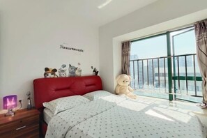 Un dormitorio con cabecera roja, un ventanal con vistas a edificios, un oso de peluche y una pared decorada con pegatinas de gatos.