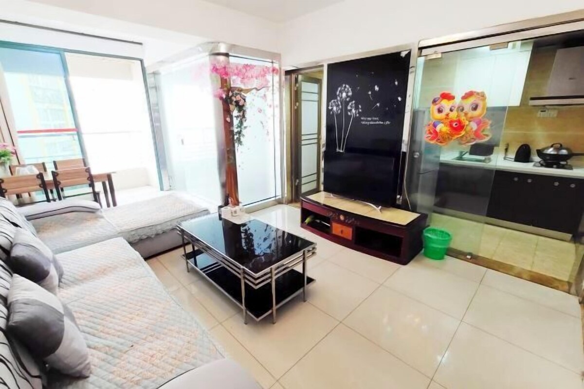 Sala de estar con sofá, una mesa de centro, televisor y una cocina visible a través de un separador de vidrio.