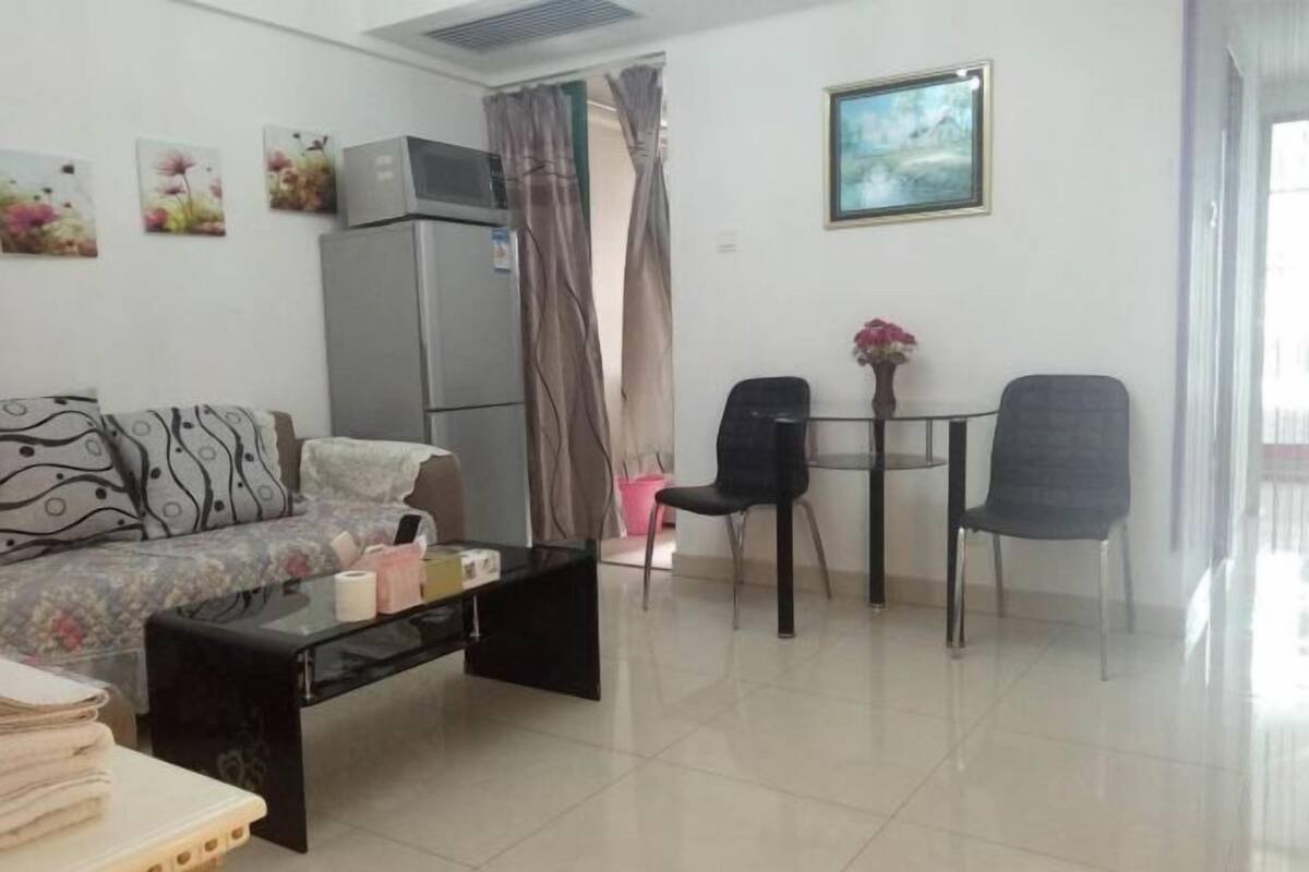 Un salon comprenant un canapé, une table basse et un coin repas avec des chaises. On y trouve également un réfrigérateur et un tableau accroché au mur.
