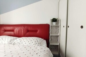 Un dormitorio con un cabecero tapizado rojo, un edredón blanco estampado y un estante blanco con una planta.