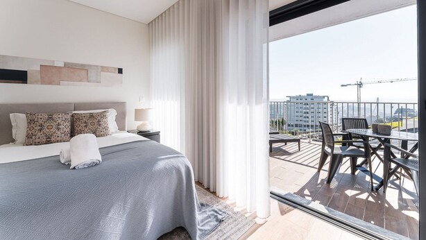 Habitación de hotel moderna con una cama grande, un balcón con mobiliario exterior y vistas a la ciudad.