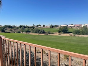 Vista de un campo de golf con cielo despejado, rodeado de vegetación escasa y edificios al fondo.