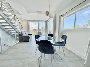 Enjoy meals at the central dining table surrounded by comfortable seating, with a staircase leading to an upper level and large windows offering views of the surrounding area.