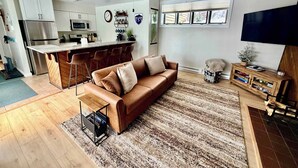 En moderne opholdsstue med sofa, sofabord, fjernsyn og et køkken med køleskab og mikroovn.