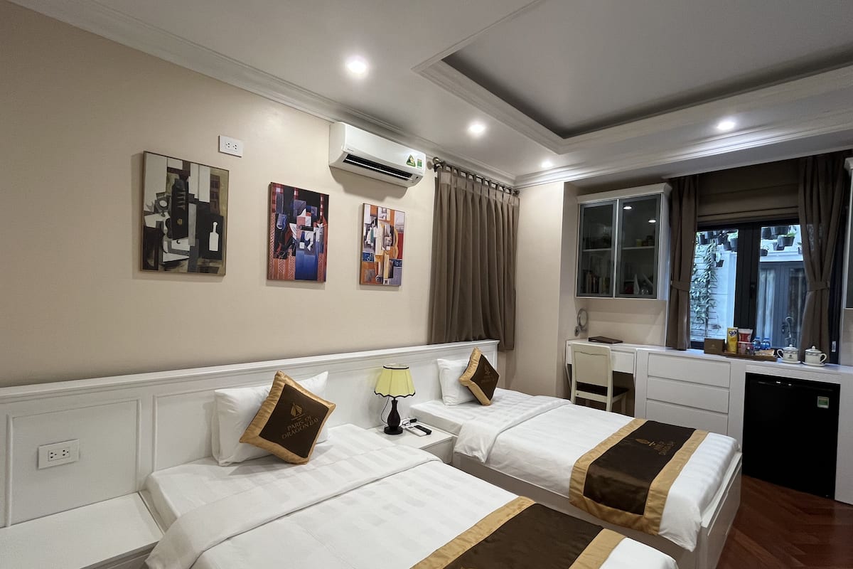 A hotel room with two beds, a desk, a window with curtains, and artwork on the wall.