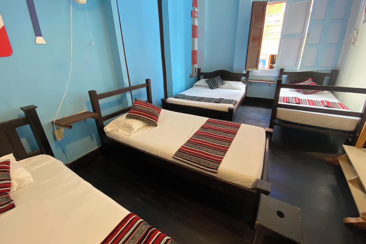 Quarto de dormitório com três camas de solteiro, cada uma com um edredom e travesseiro listrados. Há um ventilador na parede e uma janela com vista para o exterior.