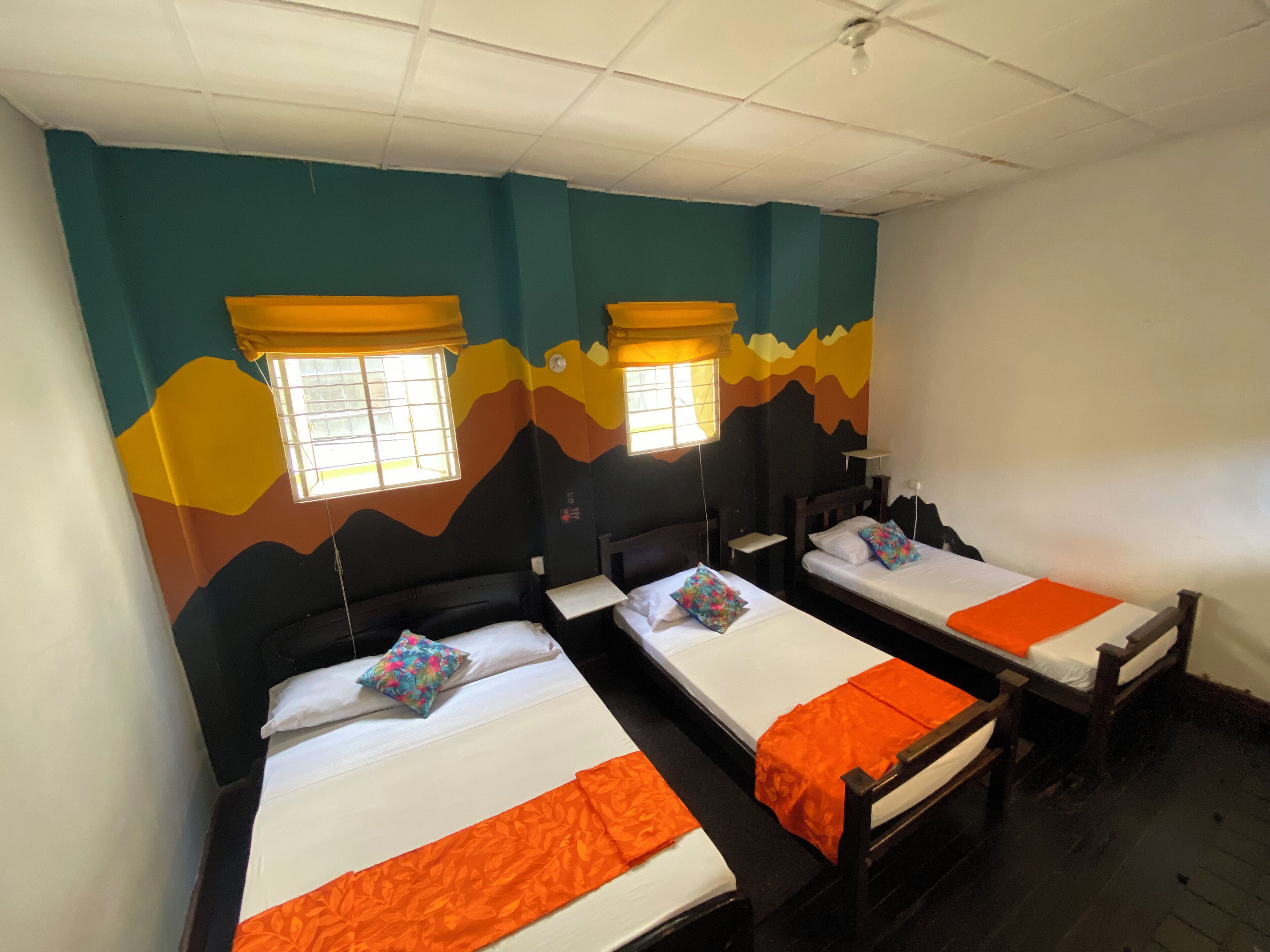 A room with three single beds, each with a white mattress and orange bedspread. The walls are painted in a gradient of blue, green, and yellow. There are windows with yellow curtains, and the room has a simple, functional design.