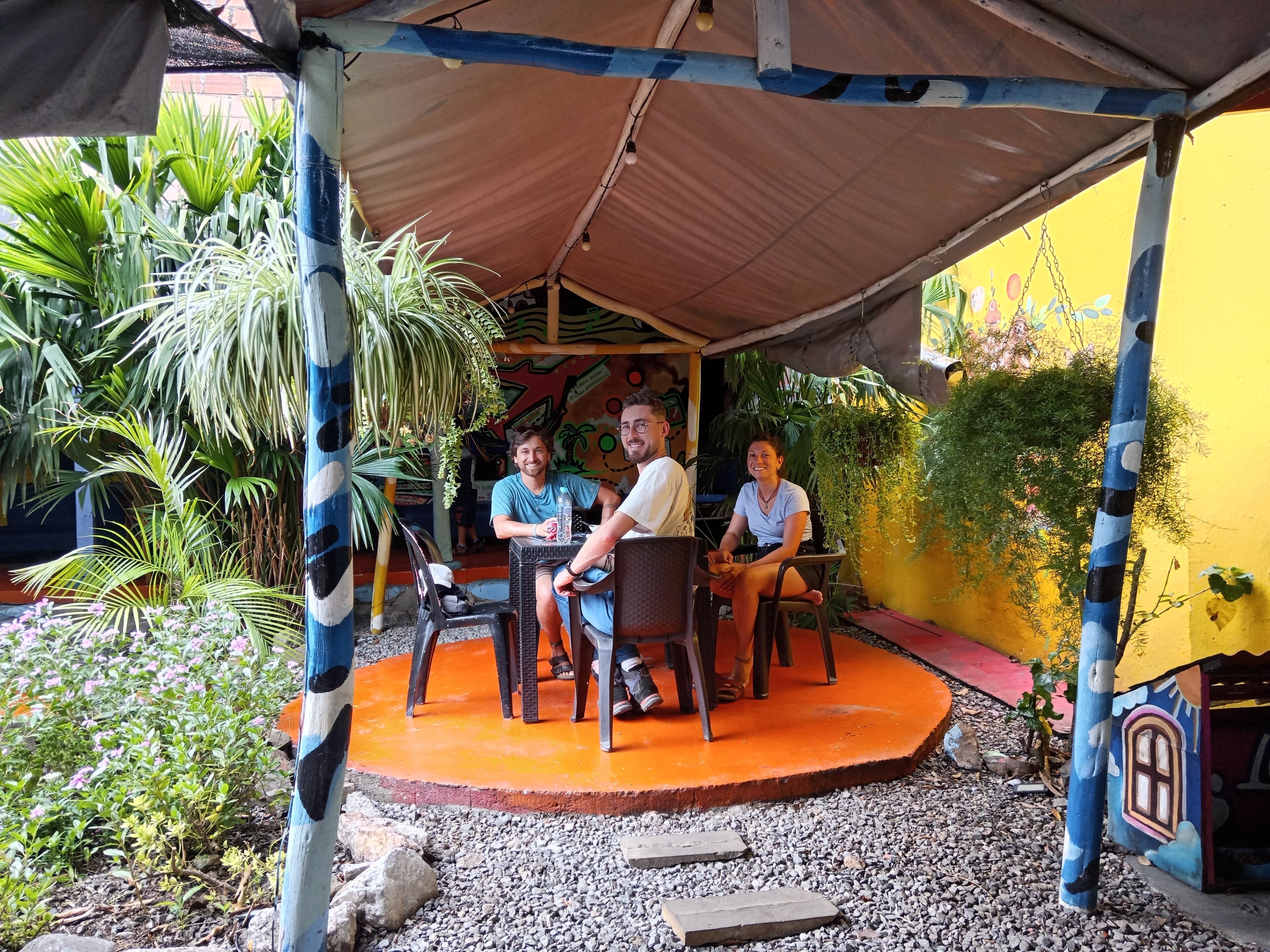 A covered outdoor seating area with a table and chairs, surrounded by plants and a colorful mural.