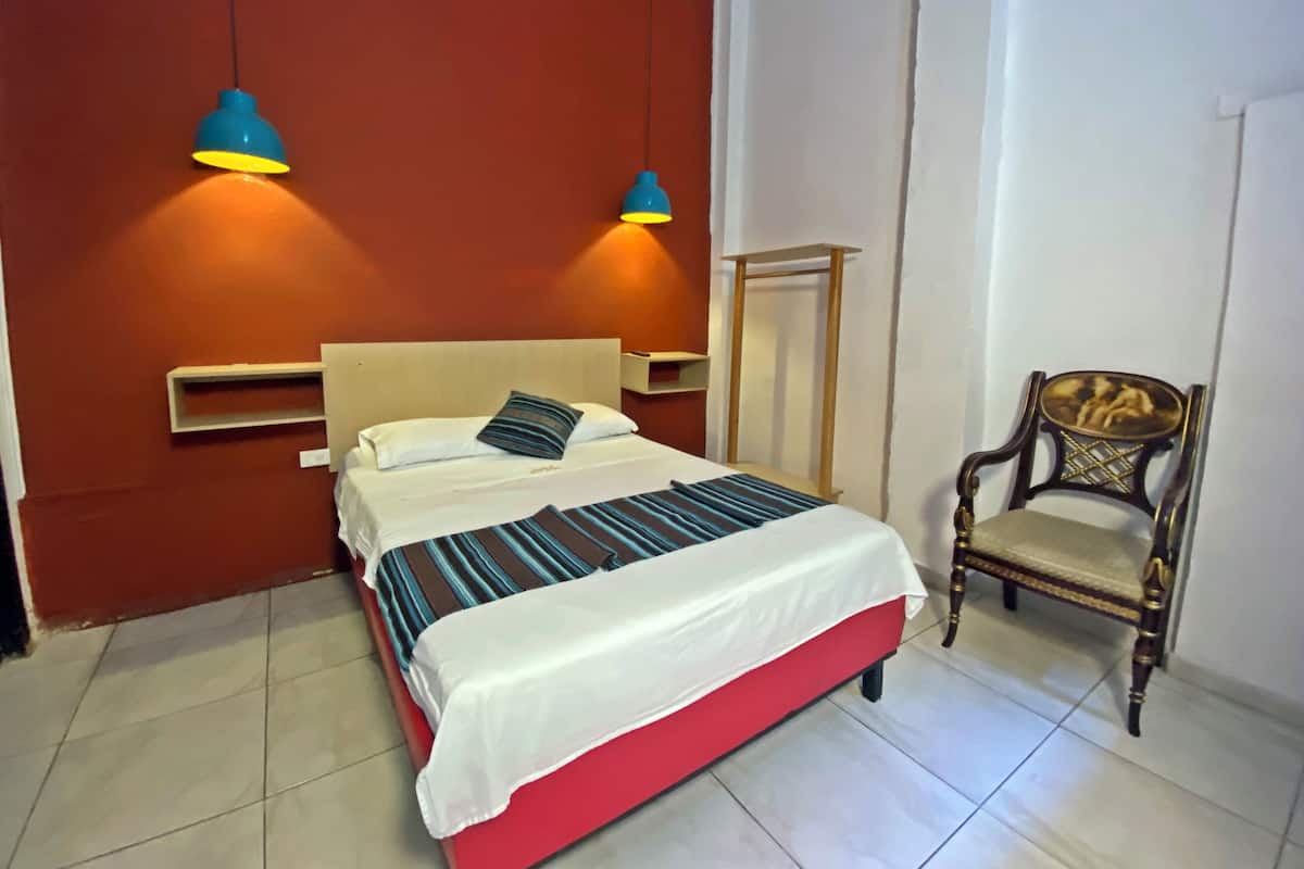 Quarto com cama vermelha, roupa de cama branca, duas luminárias na parede, uma cadeira de madeira e uma pequena prateleira.