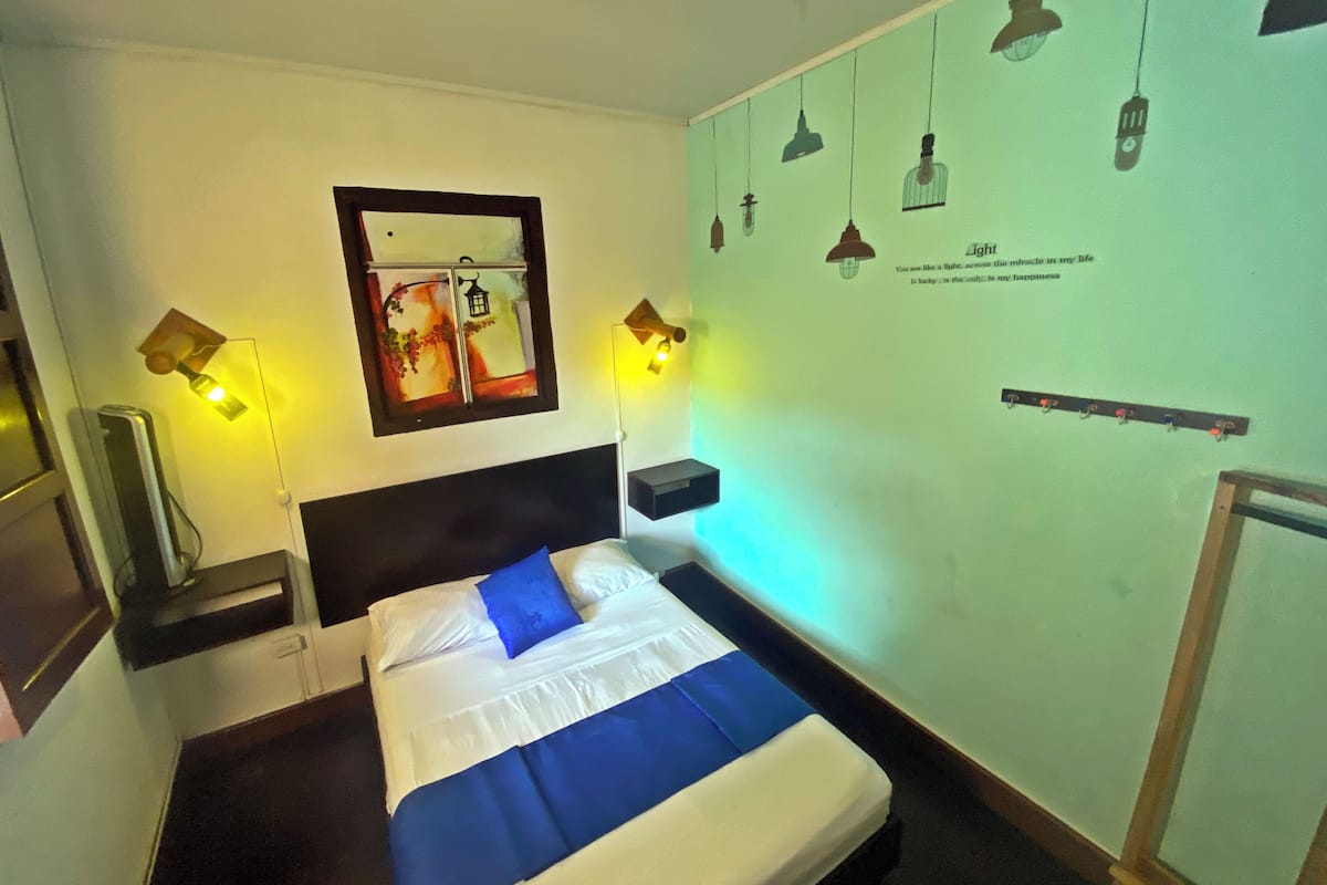 Quarto de hotel com cama, abajures, mesa de cabeceira, espelho e uma parede com luzes pendentes e uma frase.