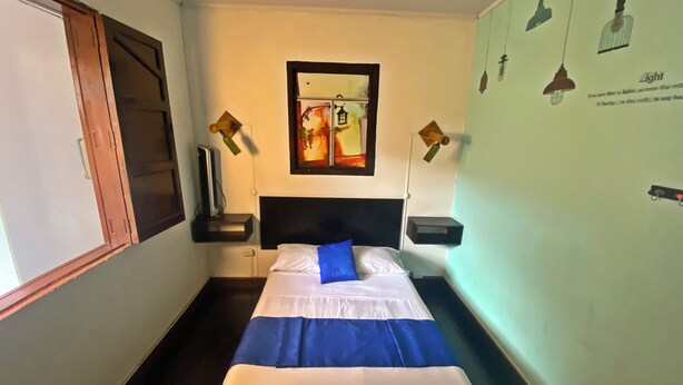 Habitación de hotel con cama, escritorio, silla, ventana y pared con cuadro e iluminación.