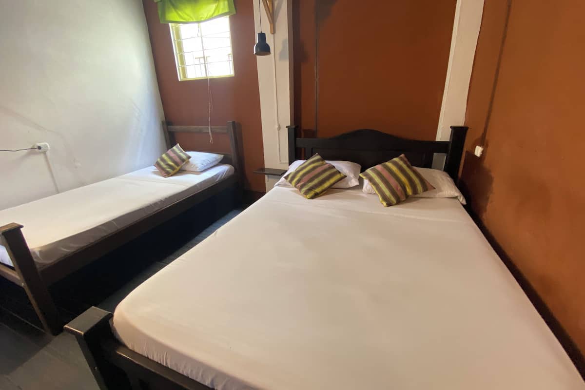 Duas camas de solteiro com estrutura de madeira, roupa de cama branca e almofadas listradas. Há uma janela com cortina verde e uma luminária pendente.