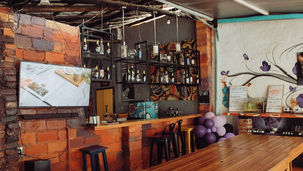 A bar area with a brick wall, wooden table, bar stools, shelves with bottles, and a mural on the wall.