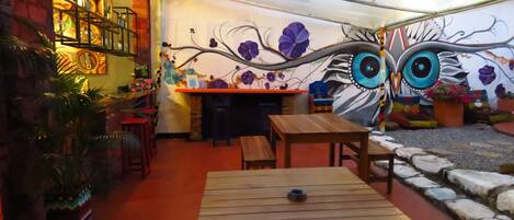 Un patio interior con mesas y bancos de madera, un mural con diseños florales y de búhos, y una pared de ladrillo con estantes.