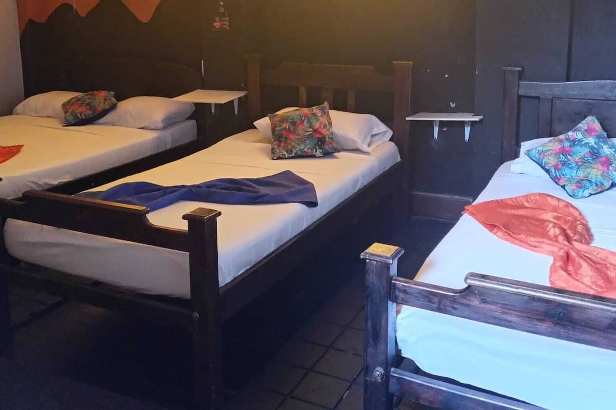 Quarto com três camas simples, cada uma com estrutura de madeira e roupa de cama branca. Há duas janelas com cortinas amarelas, e as paredes estão pintadas com um degradê de castanho e laranja.