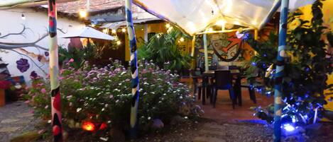 A covered outdoor area with a table and chairs, surrounded by plants and a mural on the wall.