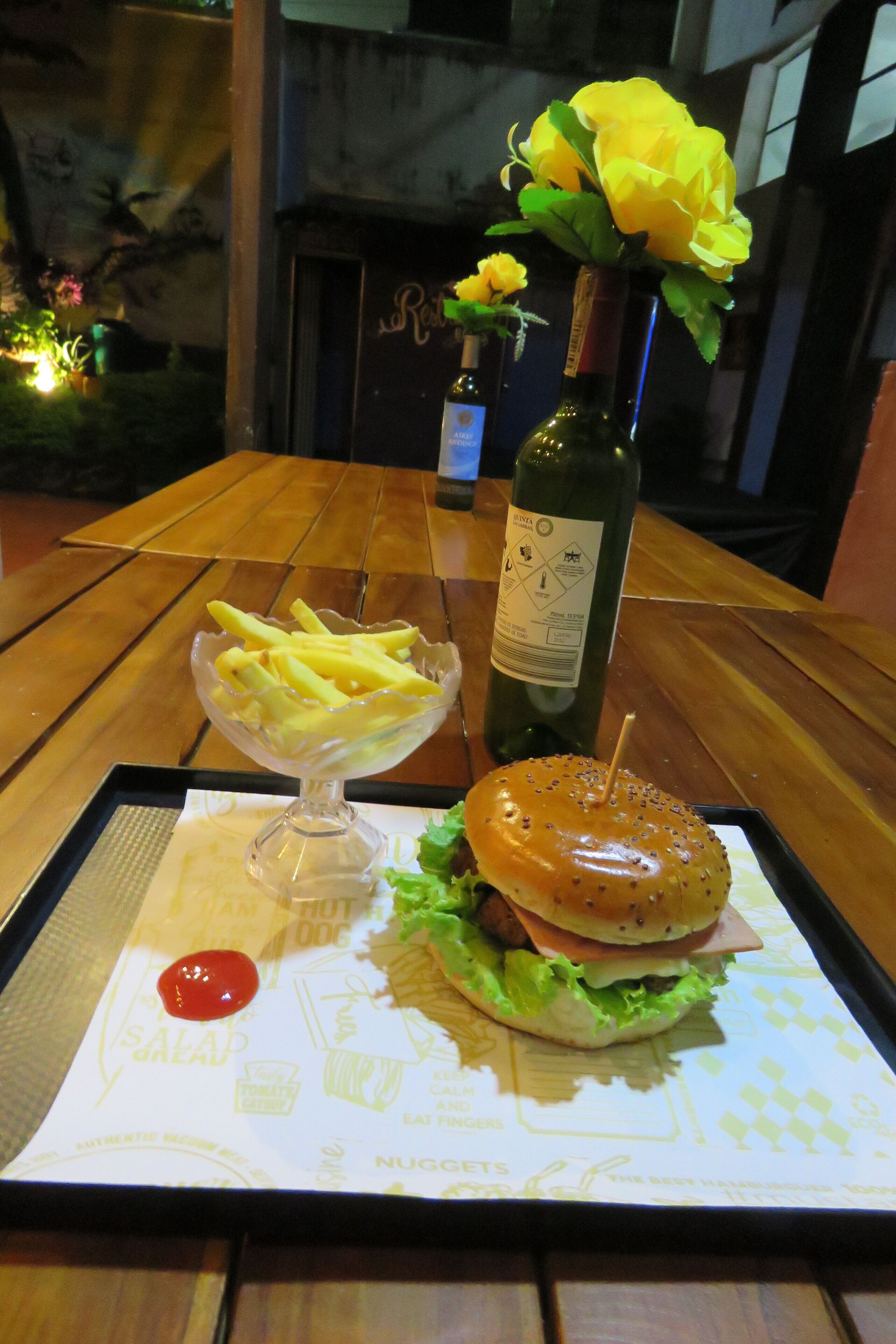 En bricka med en hamburgare, pommes frites och en sallad, tillsammans med en flaska vin och en vas med gula blommor.