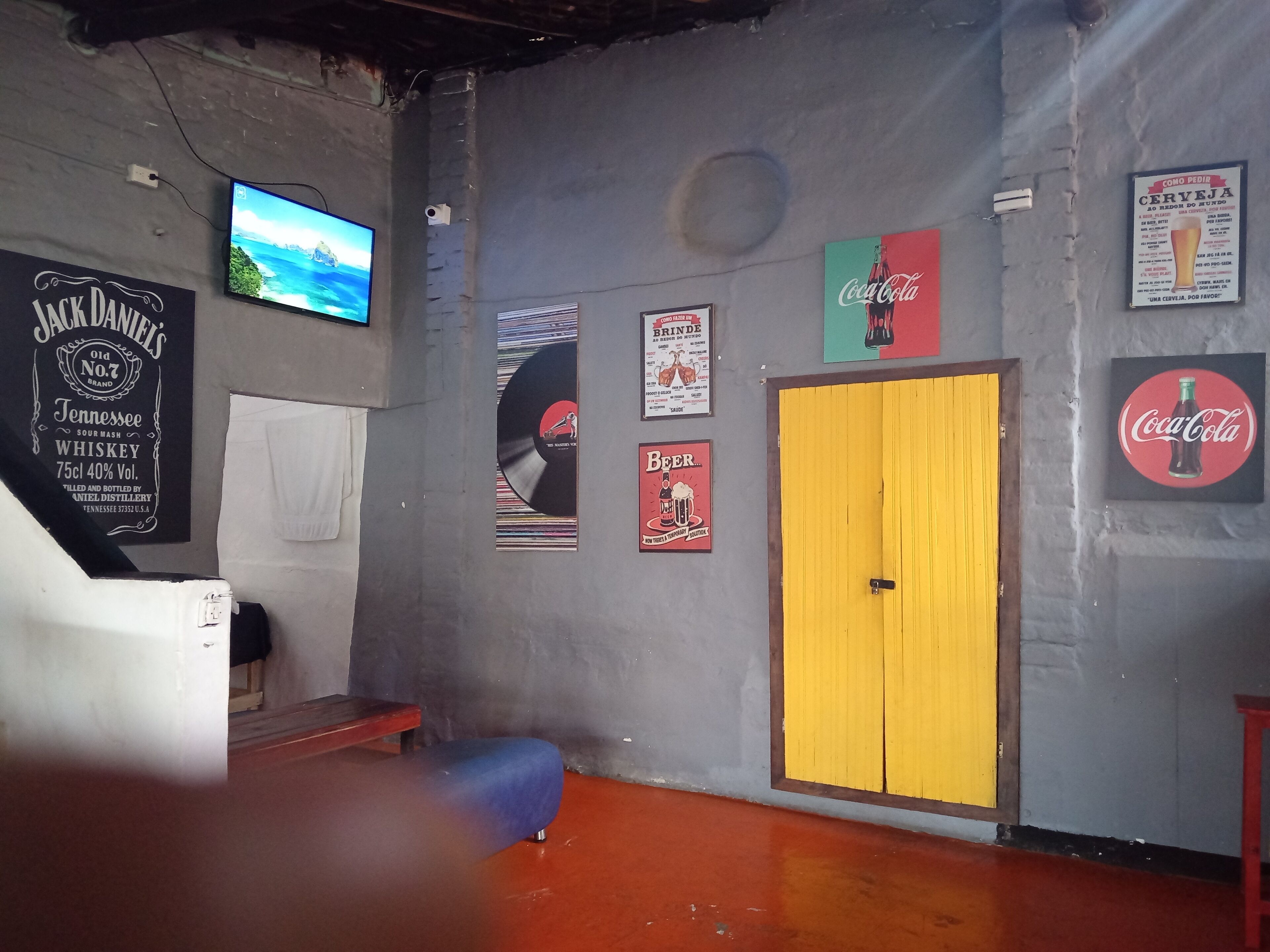 Quarto com televisão, cartazes, porta amarela e um banco vermelho.