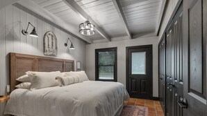 A bedroom with a large bed, a wooden headboard, a nightstand, a wall-mounted lamp, a door with a window, and a patterned rug.