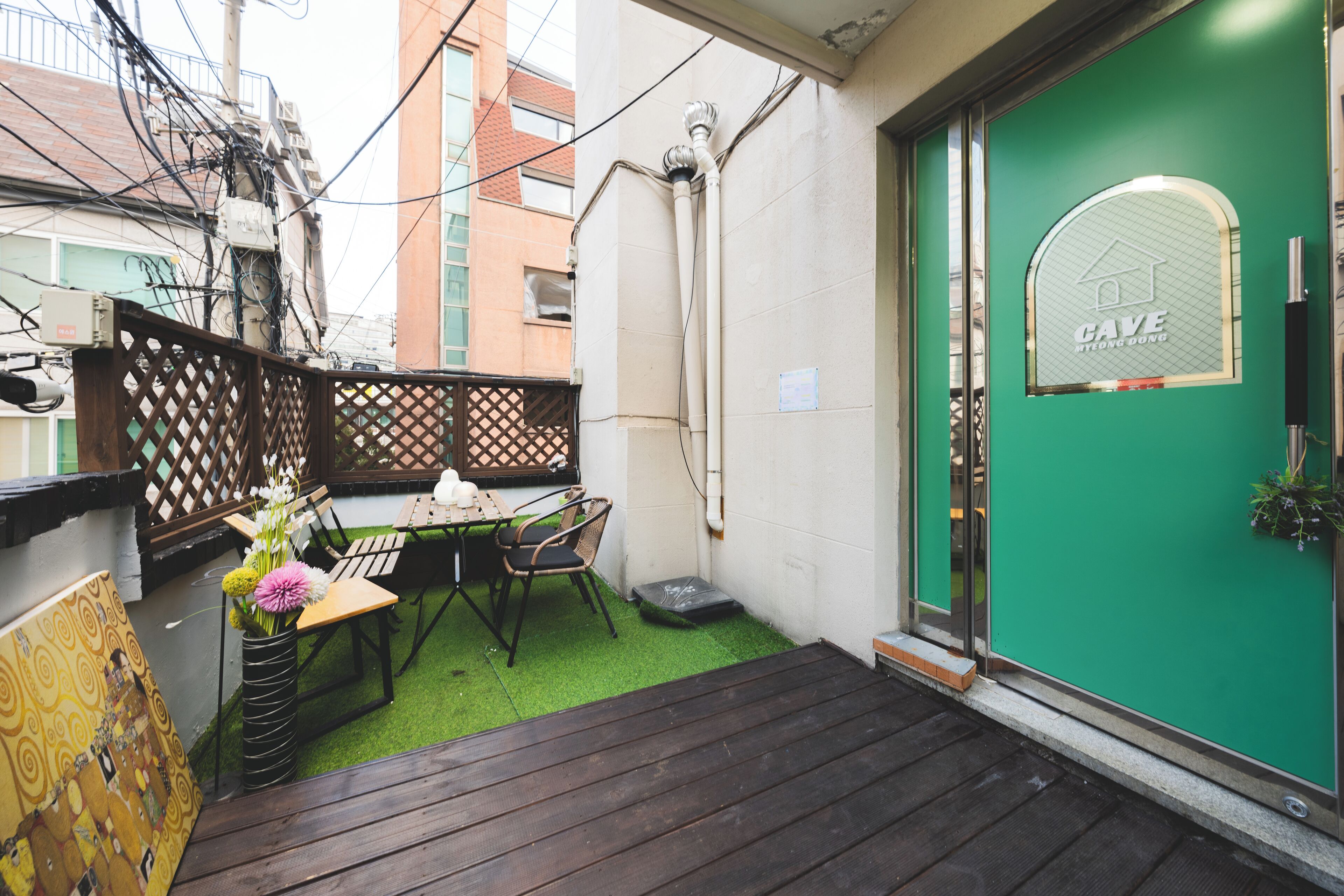 Un pequeño patio exterior con una mesa y sillas de madera, una puerta verde con un ventanal y un jardín.