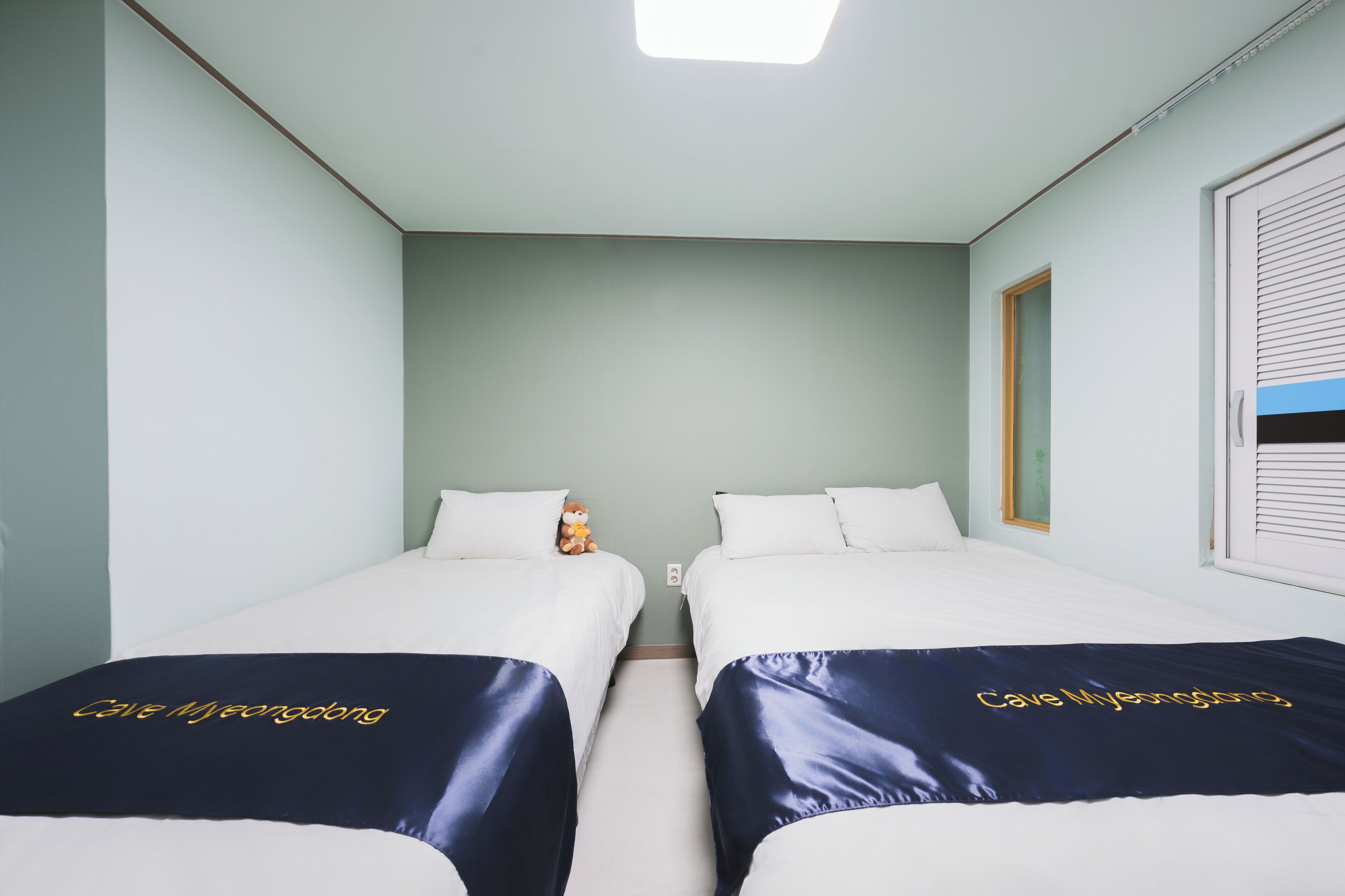 A hotel room with two single beds, each with a dark blue bedspread and the name "Cave Hjønengborg" embroidered on it. The room has light green walls, a white ceiling, and a window with blinds.