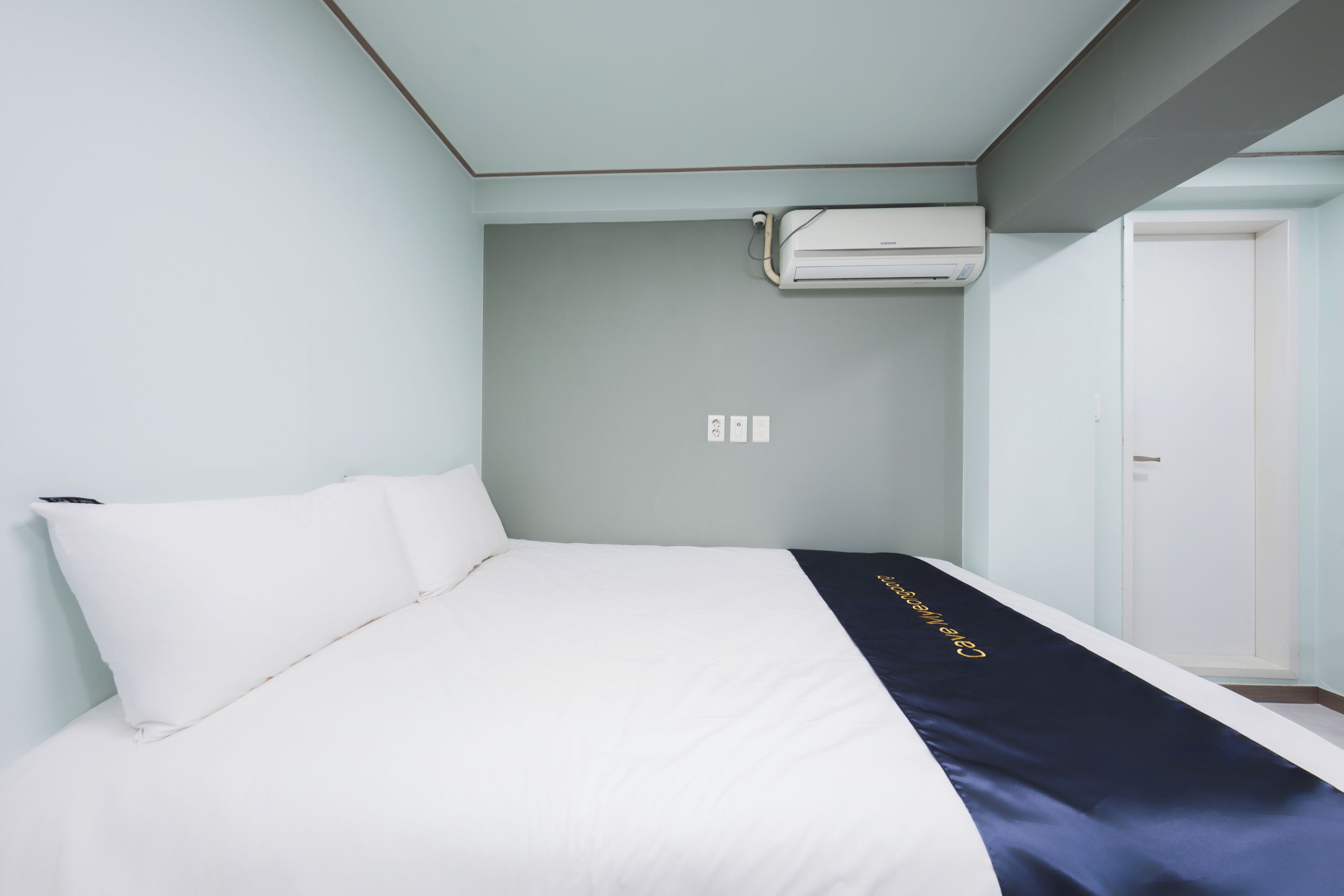 A hotel room with a bed, a bedside table, an air conditioning unit, and a door.