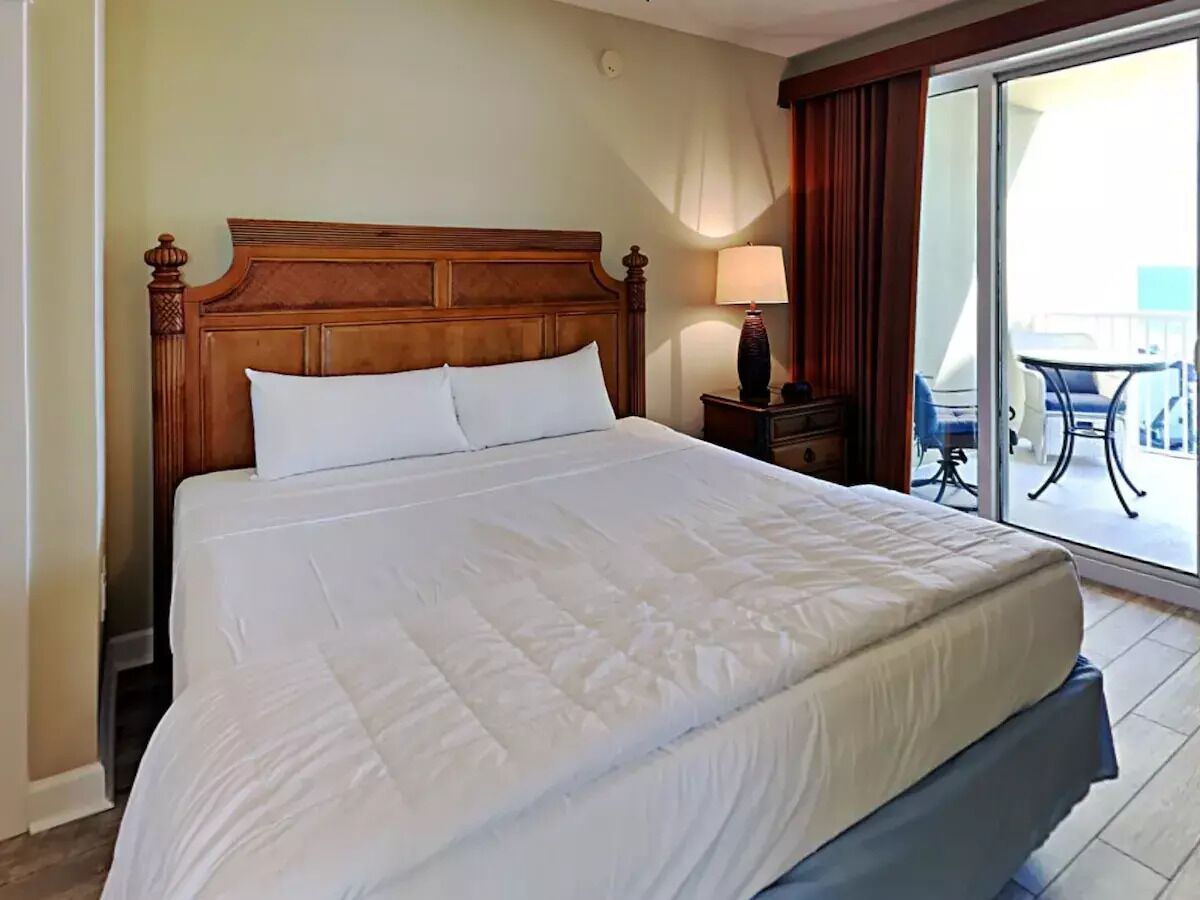 Una cama grande con cabecera de madera, una mesita de noche con lámpara y vistas a un balcón exterior.