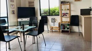 Un moderno salón con una mesa de comedor de cristal, sillas negras, un televisor de pantalla plana, una estantería y un ventanal con vistas a la vegetación.