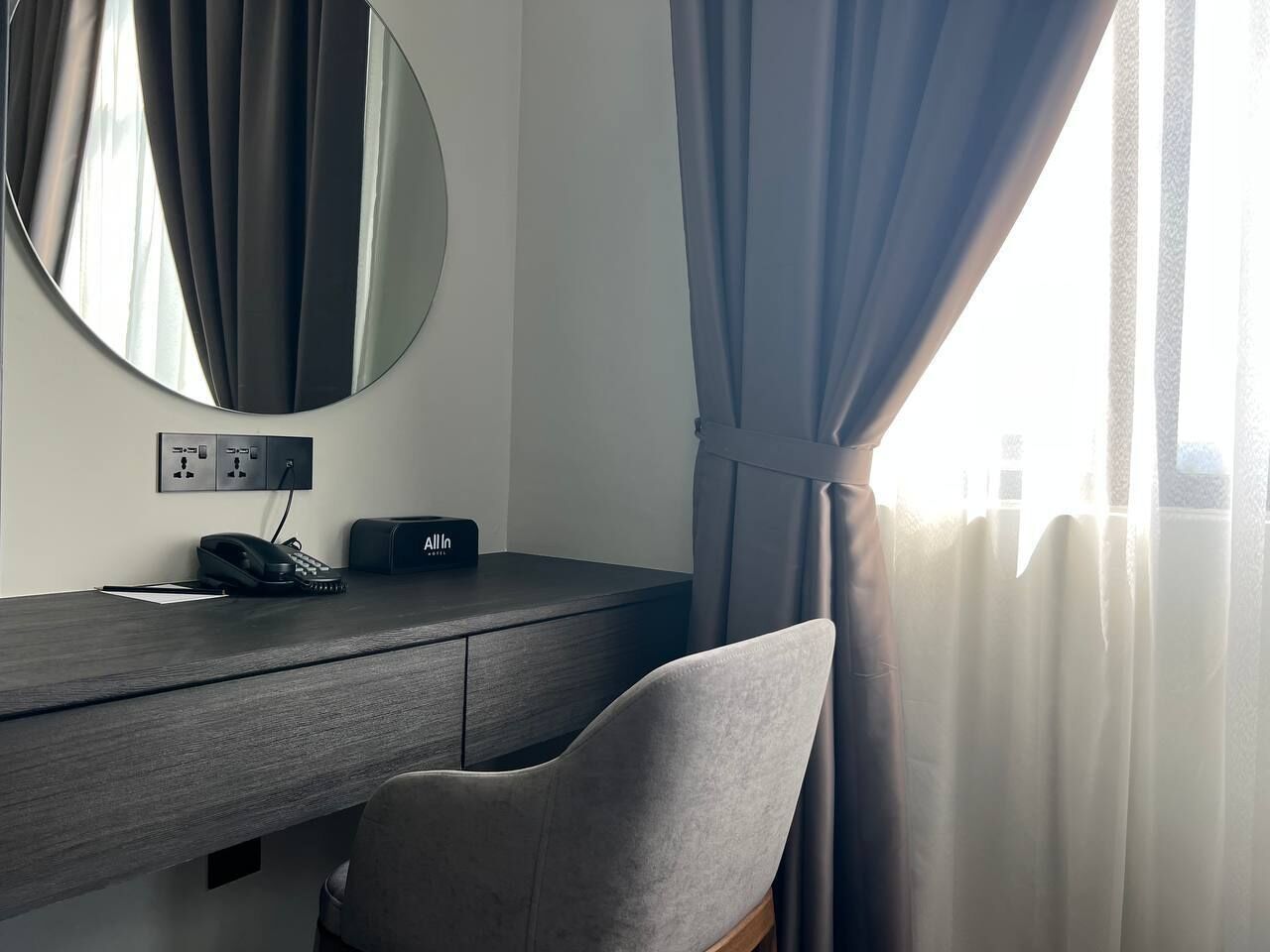 A hotel room with a desk, a chair, a telephone, and a power outlet.
