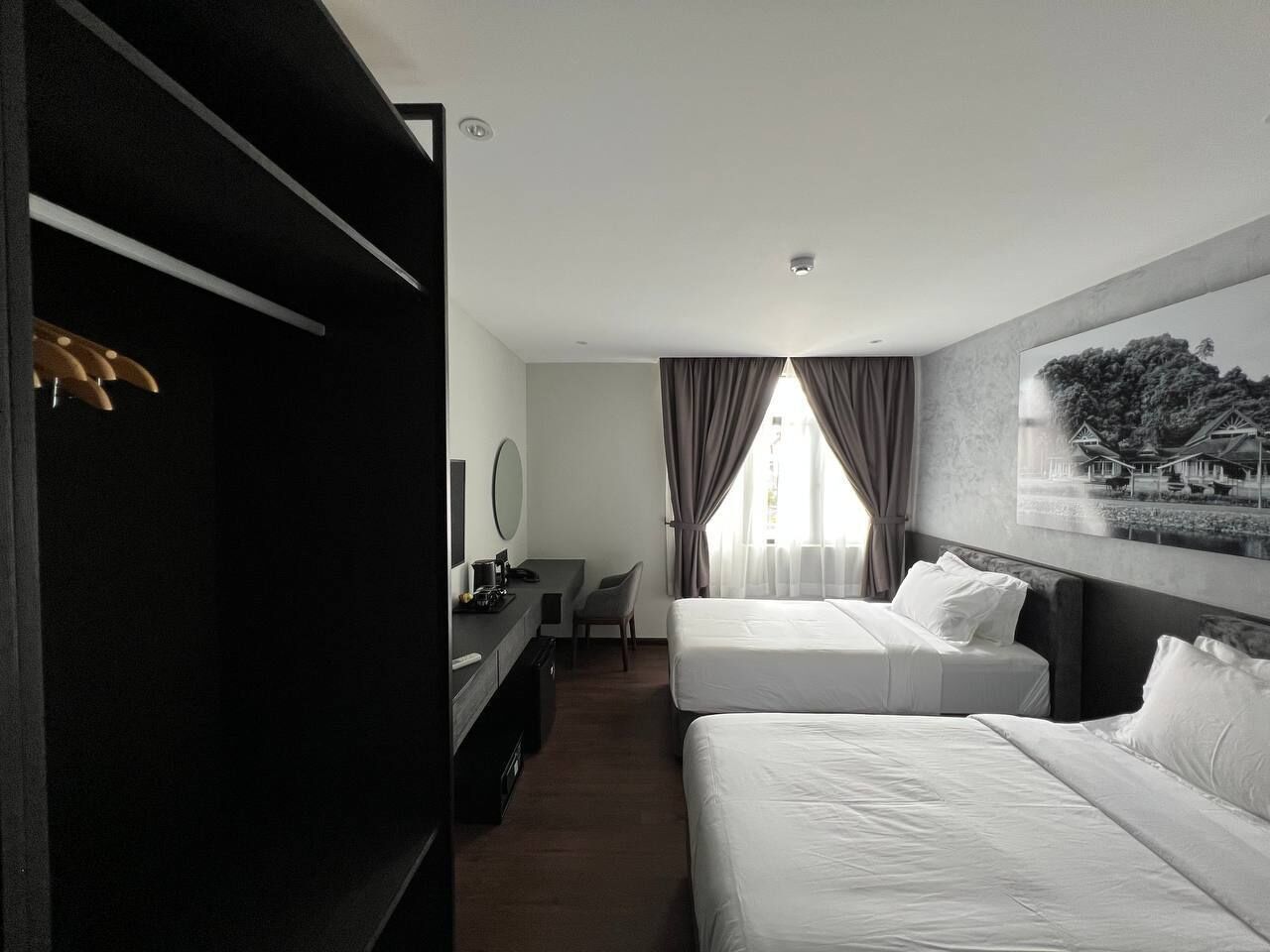 A hotel room with two beds, a desk, a chair, a coat rack, and a painting on the wall.