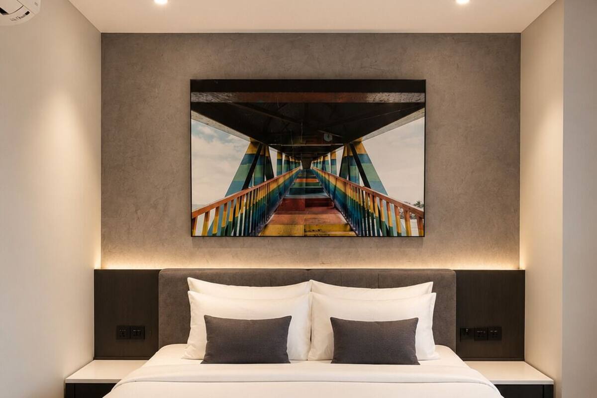 A modern hotel room with a large bed, a headboard with integrated lighting, a nightstand, and a wall-mounted artwork depicting a colorful bridge.