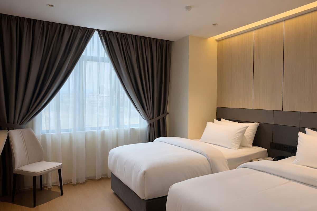 A hotel room with two beds, a chair, a large window with curtains, and a wooden headboard.