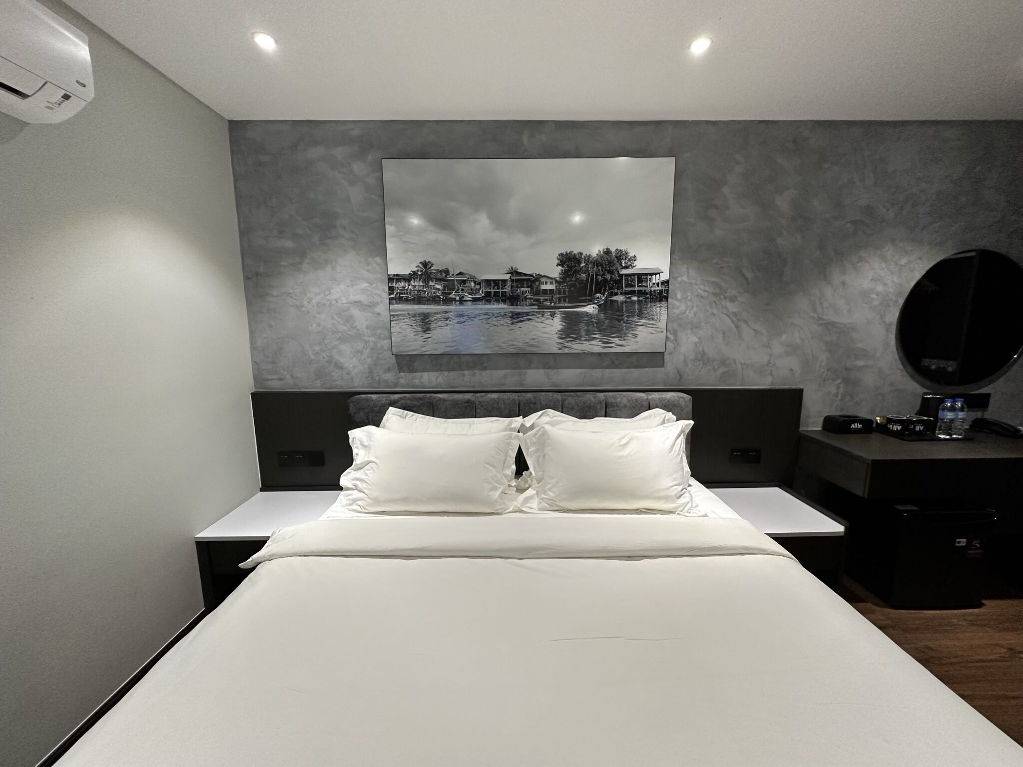 A modern hotel room with a large bed, a flat-screen TV mounted on the wall, a bedside table with a lamp, and a small round mirror.