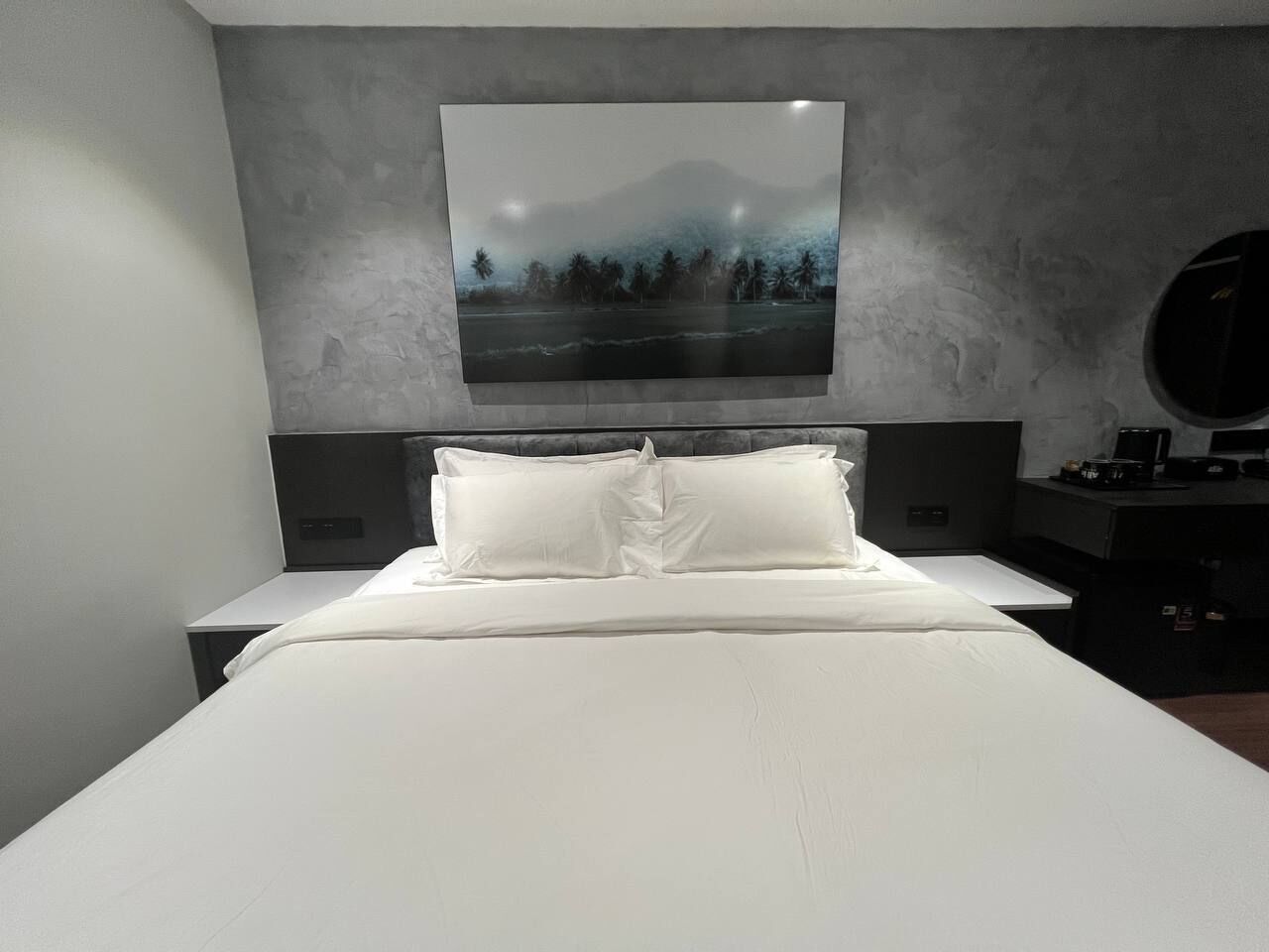 Ein modernes Hotelzimmer mit einem großen Bett, einem Nachttisch, einem runden Spiegel und einem an der Wand hängenden Landschaftsbild.