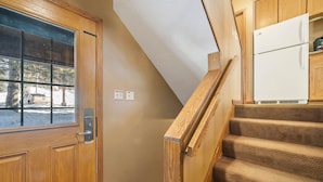 Una puerta de madera con un ventanal, una escalera con barandillas de madera y un refrigerador blanco en una zona de cocina.