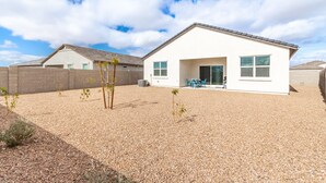 Une maison moderne de plain-pied, avec une allée gravillonnée, un petit patio doté de chaises bleues et un ciel d’un bleu éclatant.