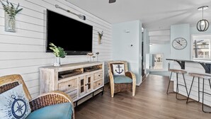 A living room with a television, wicker furniture, a bar area, and a clock on the wall. - Family Tides: Waves, Sun and PrimeTime fun for EvERYONE! (Carolina Beach)