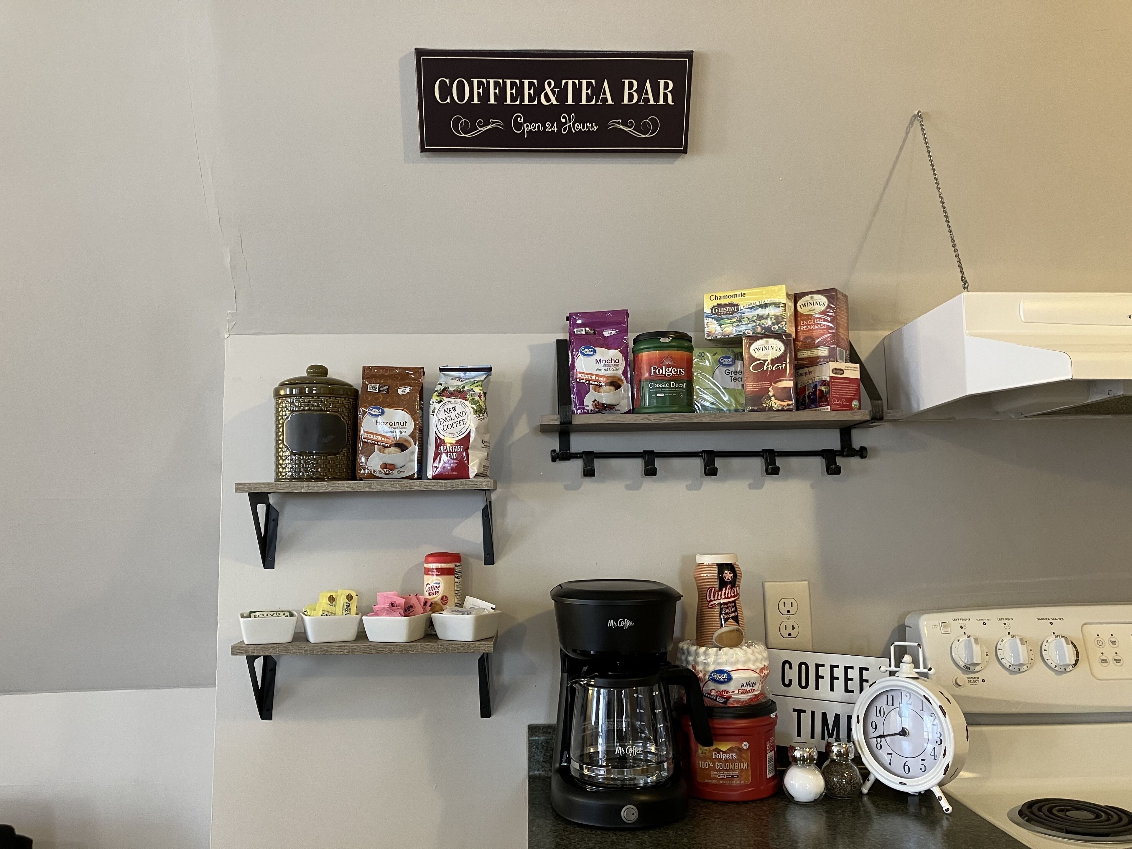A kitchen area with a coffee and tea bar sign, shelves with coffee products, a coffee maker, and a clock.
