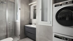 A modern bathroom with a glass-enclosed shower, a sink with a mirror, and a stacked washer and dryer. - Beachfront Elegance with Spectacular Sea Views in Kavouri Vouliagmeni  (Vouliagmeni)