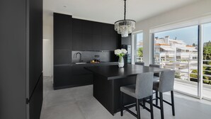 A modern kitchen with a dark island, a chandelier, and a view of buildings through large windows. - Beachfront Elegance with Spectacular Sea Views in Kavouri Vouliagmeni  (Vouliagmeni)