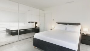 A modern bedroom with a large mirror, a bed with white linens, a dark headboard, and a nightstand with a lamp. - Beachfront Elegance with Spectacular Sea Views in Kavouri Vouliagmeni  (Vouliagmeni)