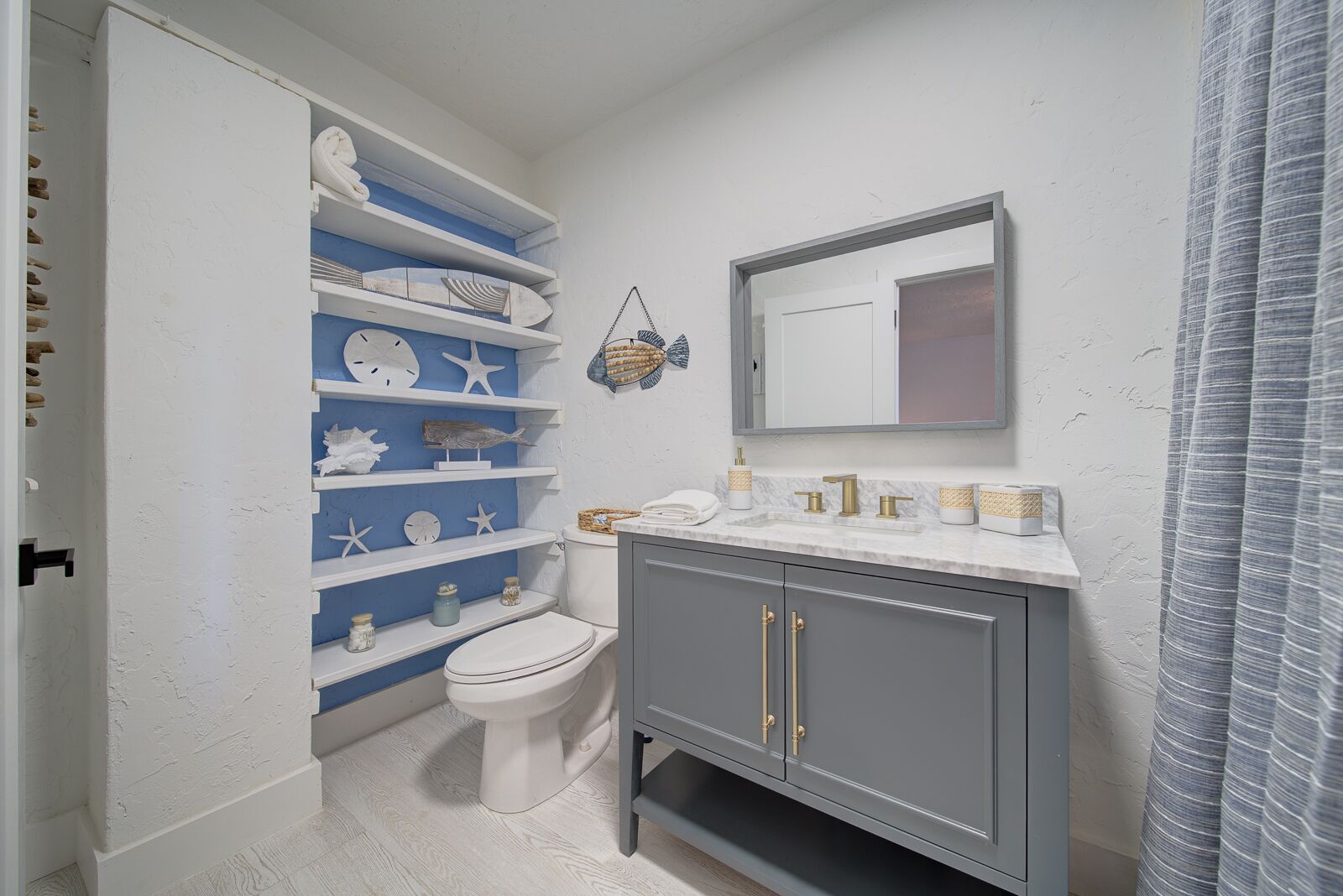 Une salle de bain avec des toilettes blanches, un meuble-lavabo avec un miroir et des étagères ornées d’objets décoratifs.