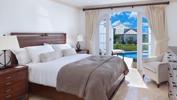 Un dormitorio con una cama grande, una mesita de noche, un aparador y vistas a una piscina y una casa.
