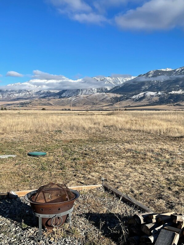 A rural landscape with a metal fire pit and logs, a green frisbee, and a clear view of snow-capped mountains under a blue sky.