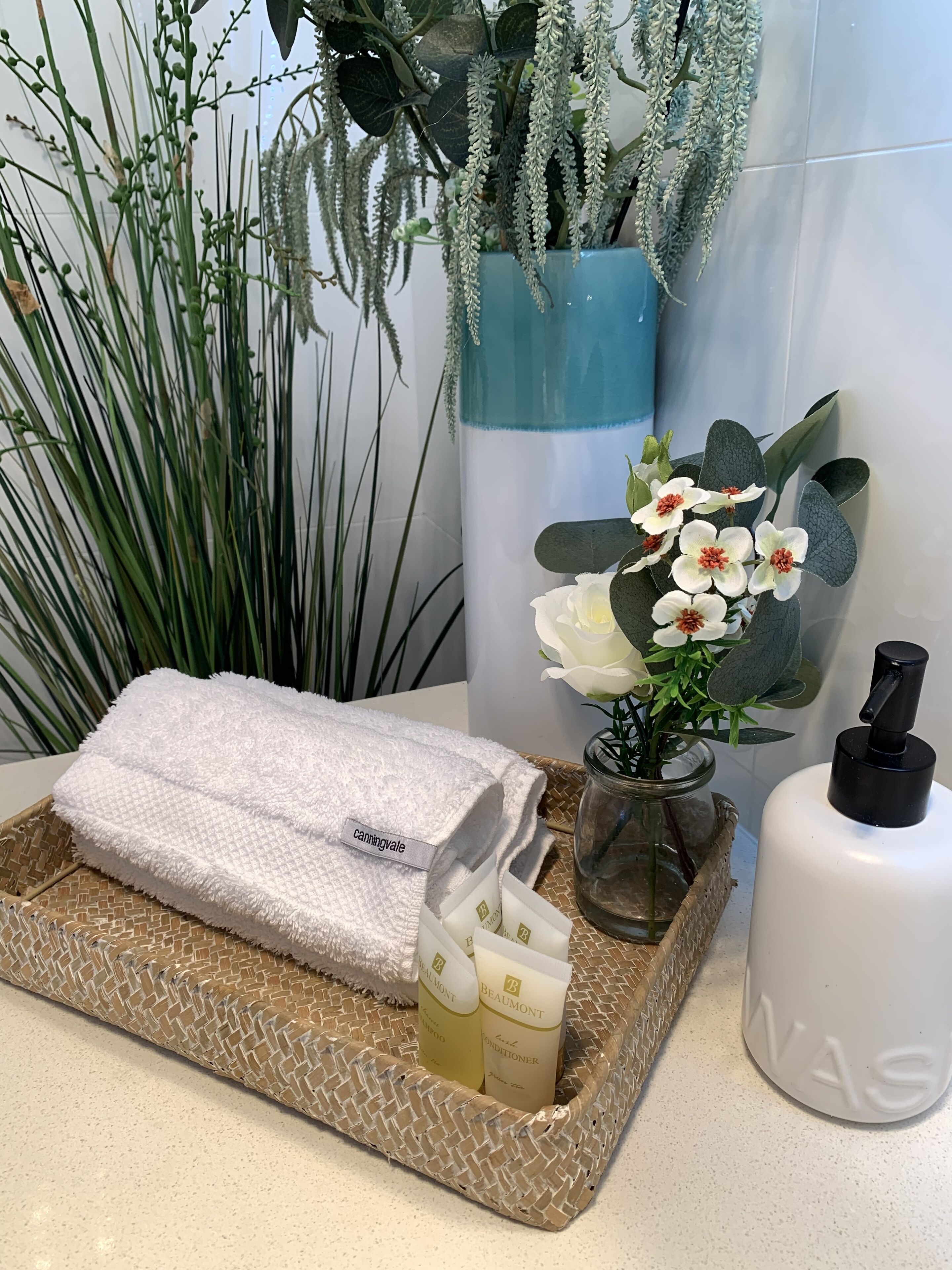 A bathroom with a white towel, a bottle of shampoo, a bottle of conditioner, and a bottle of lotion on a woven tray.