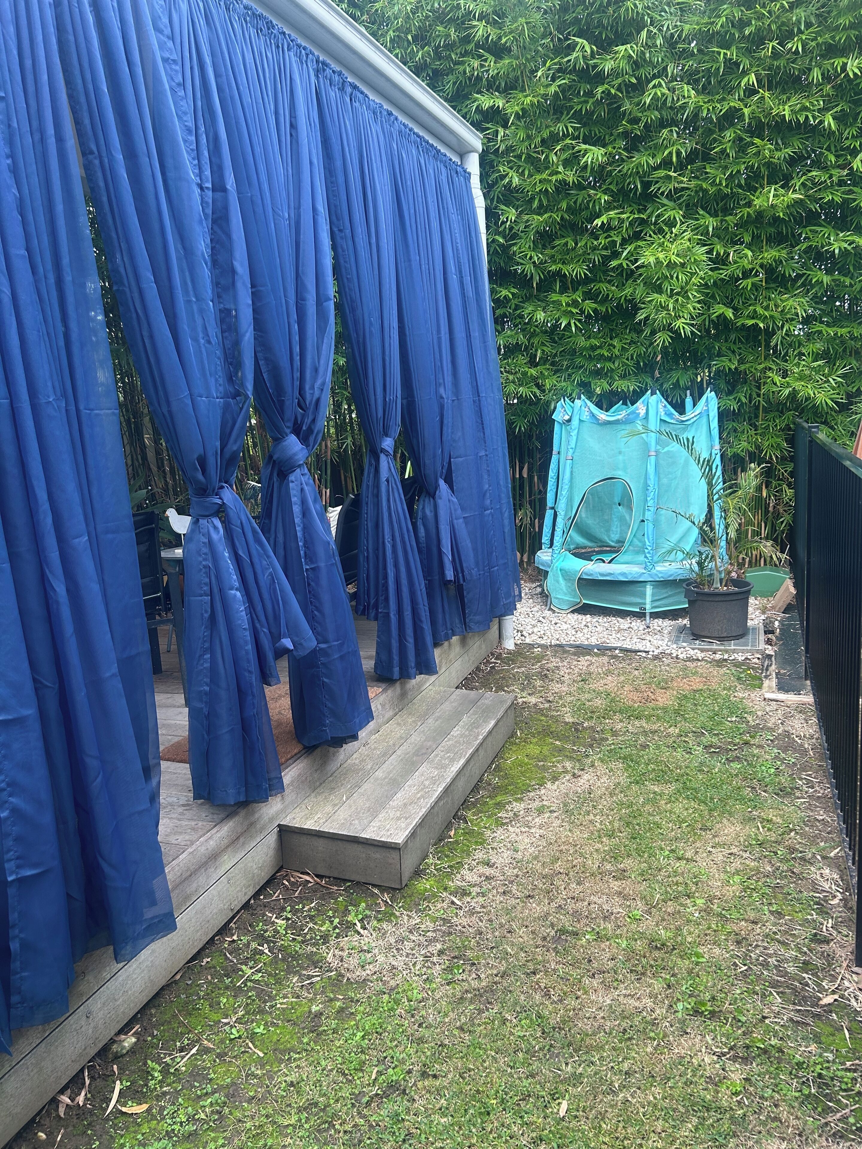 Un patio trasero con una terraza de madera, una estructura cubierta por una cortina azul y una tienda de juegos verde.