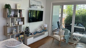 Una sala de estar con televisor de pantalla plana, estanterías de madera con libros y adornos, un pequeño árbol de Navidad, y vista a un área exterior por medio de una puerta corrediza de vidrio.