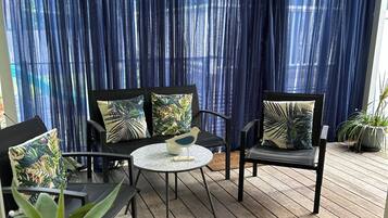Un espace extérieur aménagé avec une zone salon couverte, comprenant une table ronde et des chaises, le tout entouré de plantes en pot et de rideaux bleus.